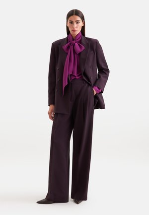 Donna che indossa un completo doppiopetto viola scuro con pantaloni a gamba larga e una camicetta viola brillante legata con un grande fiocco al collo.