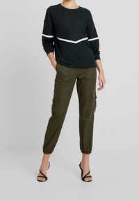 Felpa nera in pizzo con accento a righe bianche e nere, abbinata a pantaloni cargo verde oliva con polsini elastici e tasche. Sandali neri con cinturini.