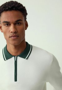 White knit polo shirt with a green collar featuring a double stripe. Zipper closure at the neckline. Smooth texture, structured fit.