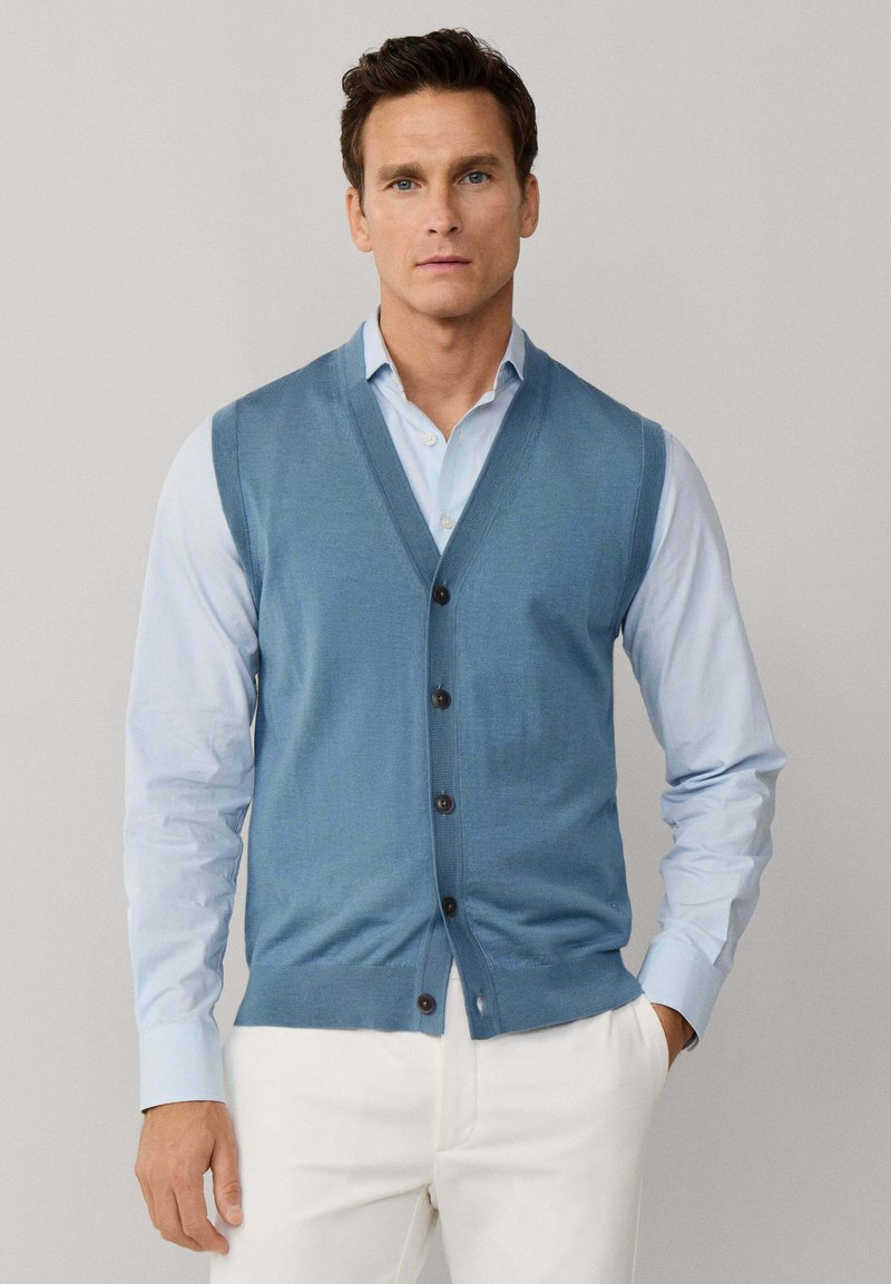 Homme portant un gilet sans manches bleu clair boutonné sur une chemise bleu pâle et un pantalon blanc, debout avec une main dans la poche contre un fond gris.