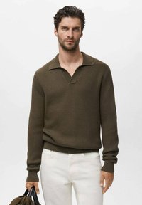 Suéter de punto acanalado verde oliva con cuello tipo polo y mangas largas, combinado con pantalones blancos. La modelo sostiene un bolso negro.