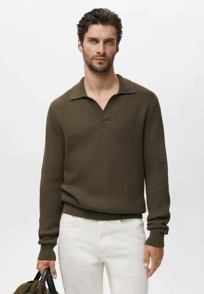 Suéter de punto acanalado verde oliva con cuello tipo polo y mangas largas, combinado con pantalones blancos. La modelo sostiene un bolso negro.