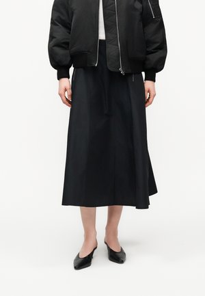 Naine, kes kannab musta bomberjopet, musta midi-seelikut ja musti teravate ninadega mule jalatseid, seisab valge tausta ees.