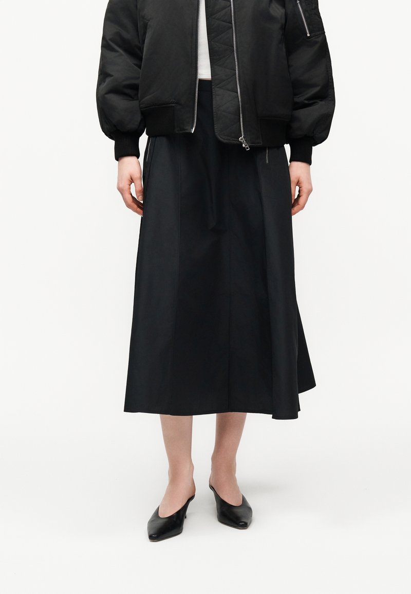 Femme portant une veste bomber noire, une jupe midi noire et des mules noires à bout pointu, debout devant un fond blanc.