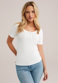 Weißes, geripptes Kurzarmoberteil mit runder Ausschnitt und fünf passenden Knöpfen auf der Vorderseite, kombiniert mit hellblauen Jeans.