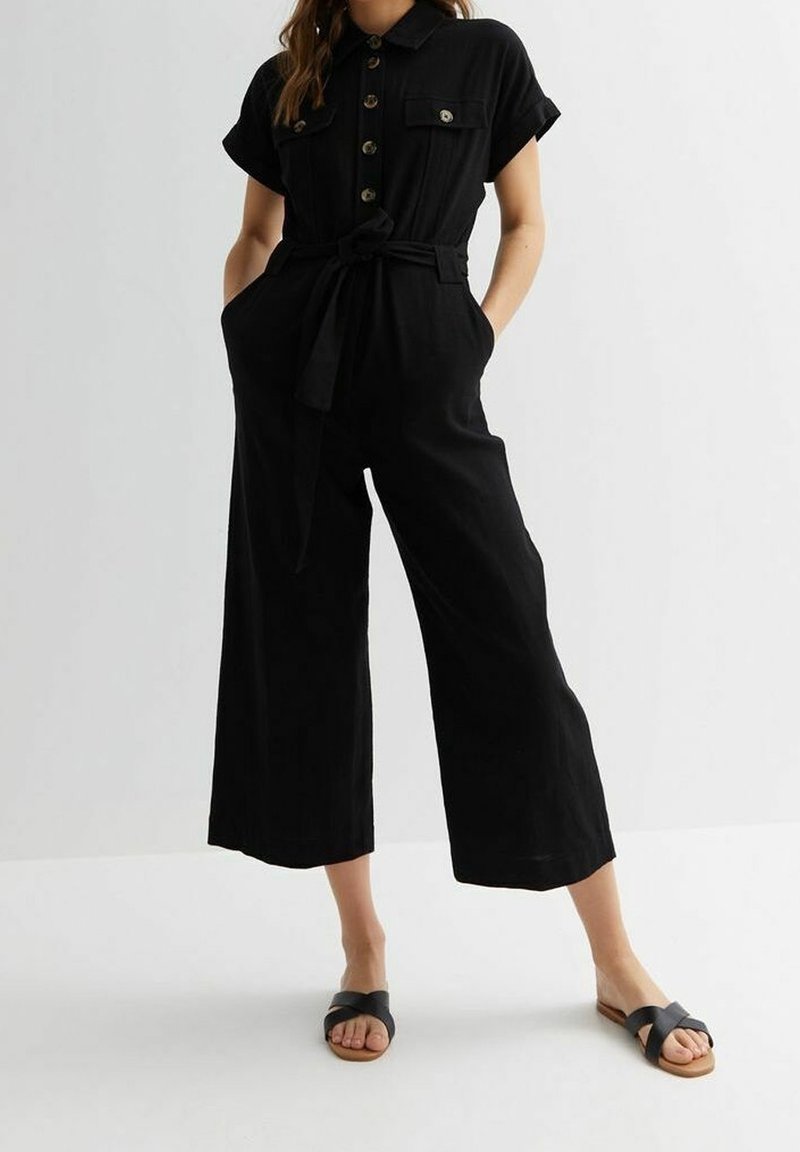 Femme portant une combinaison noire à boutons avec manches courtes, taille ceinturée, jambes larges et raccourcies, et sandales noires à enfiler, debout les mains dans les poches.