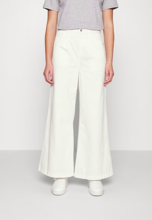 Persoon draagt witte broek met wijde pijpen, een lichtgrijs kort mouwen overhemd ingestopt en witte sneakers, staand tegen een effen lichte achtergrond.