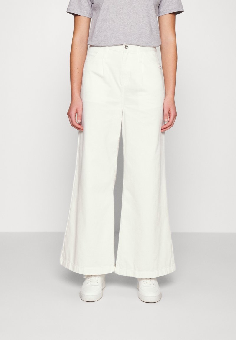 Personne portant un pantalon blanc à jambes larges, une chemise gris clair à manches courtes rentrée et des baskets blanches, debout devant un fond clair uni.