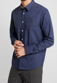 Camicia button-up blu navy con un sottile motivo geometrico, maniche lunghe e bottoni bianchi. Tessuto liscio con un design adatto.