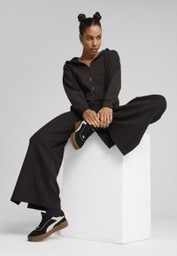 Black zip-up hoodie paired with wide-leg black pants. Model wears black and white sneakers with a textured sole. Standing on a white pedestal.