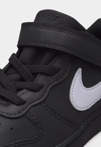 Μαύρο αθλητικό παπούτσι Nike με μαύρα κορδόνια, λευκό λογότυπο swoosh με ραφτή ραφή και μαύρο ιμάντα βέλκρο στην κορυφή.