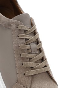 Zbliżenie na tenisówkę z zamszu w kolorze taupe i gładkiej skóry, z płaskimi plecionymi sznurowadłami przechodzącymi przez sześć oczek z każdej strony.