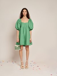 Mulher vestindo um vestido verde de mangas bufantes, segurando uma bolsa de pérolas verde e branca, em pé sobre um chão coberto de confetes coloridos.