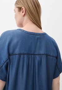 Denim top with short sleeves, deep blue color, relaxed fit, gathered stitching at back, and a rounded neckline. Smooth texture.