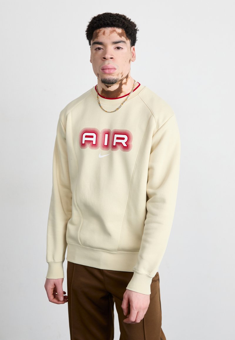 Beiger Sweatshirt mit rotem "AIR"-Logo, Rundhalsausschnitt und gerippten Bündchen. Kombiniert mit braunen Hosen, die eine subtile Textur aufweisen.