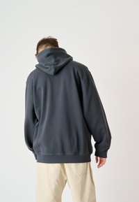 Dunkelgrauer Kapuzenpullover mit lockerer Passform. Weicher Stoff, gerippte Bündchen und Saum, große Kapuze und flache Nähte, die auf der Rückseite sichtbar sind.
