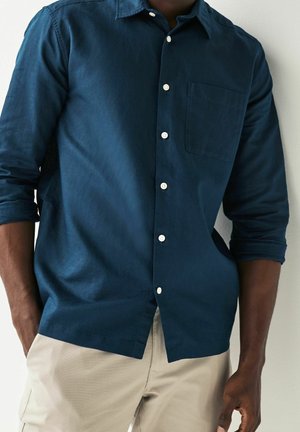 Homme portant une chemise bleue foncée à boutons avec les manches retroussées et un pantalon beige, debout avec une main dans la poche.