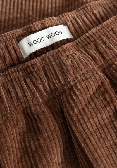 Tissu en velours côtelé marron avec des côtes verticales, équipé d'une taille élastique et d'une étiquette tissée affichant "WOOD WOOD." Texture douce.