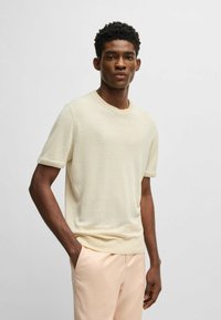 Kurzärmliger cremefarbener Strickpullover mit Rundhalsausschnitt und gerippten Säumen, kombiniert mit hellpfirsichfarbenen Hosen, präsentiert vor einem schlichten Hintergrund.
