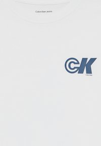 Balta medvilninė marškinėliai su tamsiai mėlyna grafika, kuri vaizduoja raides "CK" su apvaliu akcentu, o žemiau dizaino yra užrašas "Calvin Klein".
