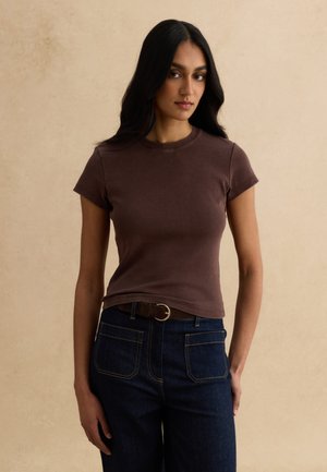 Giovane donna con lunghi capelli scuri che indossa una maglietta marrone aderente a maniche corte e jeans blu scuro a vita alta con una cintura marrone.