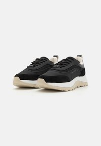 Sneaker atletiche nere realizzate in mesh e suede, con una suola beige chiaro, dettagli testurizzati e un'etichetta sul retro.