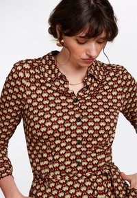 Camicia con bottoni in tessuto morbido, caratterizzata da un motivo geometrico in rosso, verde e crema. Presenta un colletto e chiusura con bottoni.