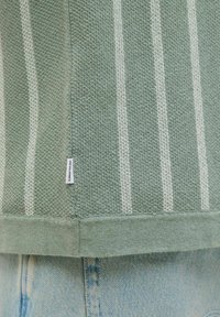 Grønstribet sweater med en struktureret vævning, der har tynde hvide striber og en ribbet kant. Inkluderer et lille mærke på sidesømmen.