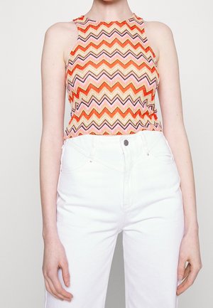 Femme portant un haut sans manches à col montant avec motif en zigzag orange, rose et beige, assorti à un pantalon blanc taille haute.