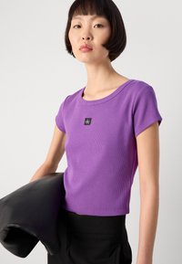 Purple ribbed short-sleeve top with a wide neckline and a small black logo patch, paired with a black leather jacket draped over the arm.