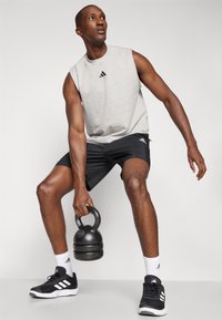Haut sans manches gris, shorts noirs, chaussures de sport noires et chaussettes blanches. Tenant un kettlebell noir. Logo Adidas visible sur les vêtements.