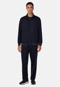 Jūrų laivyno sportinis kostiumas su užtrauktuku užsegama striuke ir atitinkančiomis kelnėmis, pasižymintis lygiu audiniu, elastingomis rankogaliais ir šoninėmis kišenėmis, derinamas su juodais sportbačiais.