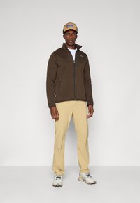 Brown fleece jacket with a zippered front, paired with tan trousers and white sneakers. A beige cap with a logo completes the outfit.