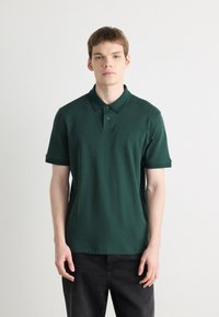 Tmavozelená polo košeľa vyrobená z hladkej bavlny, s klasickým golierom, dvomi gombíkmi na zapínanie a vyšitým logom na ľavej hrudi.