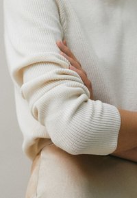 Persoon in een crèmewitte gebreide trui met opgerolde mouwen, armen over hun torso gekruist, met zichtbare beige broek.