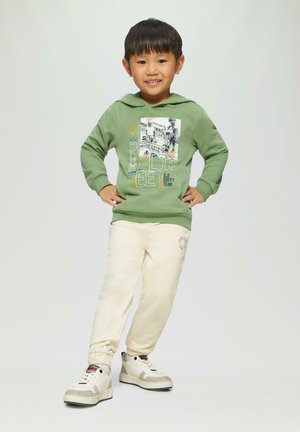 Felpa verde con testo e grafica colorati, pantaloni della tuta crema e sneakers bianche con accenti grigi, bambino in piedi con le mani sui fianchi.