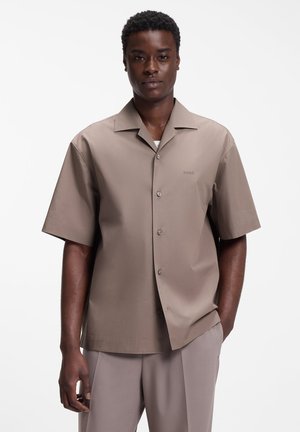 Homme portant une chemise à manches courtes taupe à boutons et un pantalon assorti, debout avec une main dans la poche devant un fond blanc.