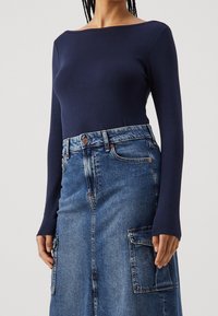 Marineblauwe langarmtop gecombineerd met eendenimrok met zakken, lichtblauwe wassing en een split aan de voorkant, die een aansluitende en gestructureerde silhouet laat zien.