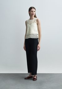 Kobieta ubrana w kremowy, bezrękawnik z szydełkowym wzorem, czarne, szerokie spodnie o fakturze oraz brązowe płaskie buty, stojąca na tle jednolitego szarego tła.