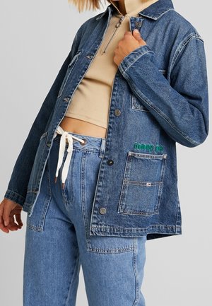 Persoon draagt lichtblauwe spijkerjas en broek met beige cropped top, houdt de kraag van de jas vast, met zichtbaar "tommy jeans" logo op het jaszakje.