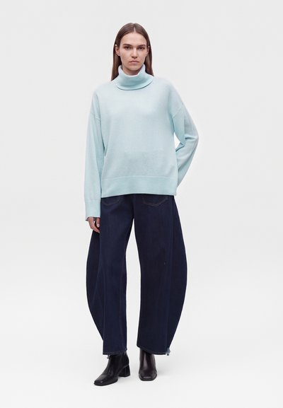 Femme portant un pull à col roulé bleu clair, un pantalon large bleu foncé et des bottines noires, debout avec une main dans la poche.