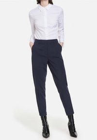 Kobieta ubrana w białą koszulę zapinaną na guziki, granatowe dopasowane spodnie i czarne szpiczaste botki za kostkę, trzymająca ręce w kieszeniach.