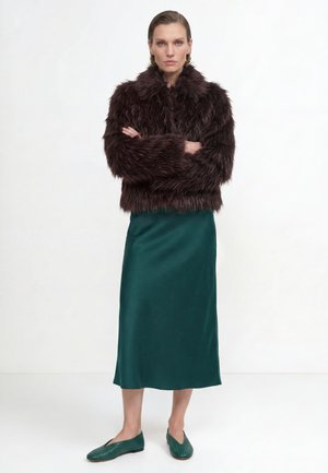 Kvinde stående med armene krydset iført en mørkebrun faux fur jakke, grøn satin nederdel, grønne flade sko og guldfarvede hoop øreringe mod hvid baggrund.