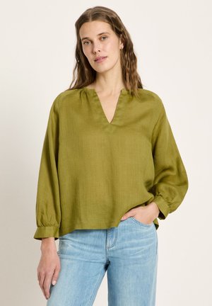 Frau mit langem, welligem Haar, trägt eine olivgrüne Bluse mit langen Ärmeln und V-Ausschnitt sowie hellblaue Jeans, eine Hand in der Tasche, neutraler Hintergrund.