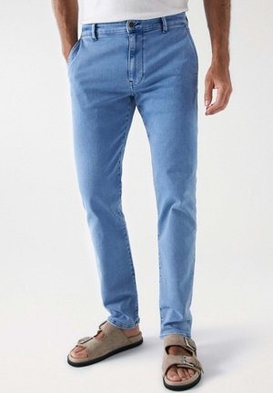 Homme portant un jean slim bleu clair et des sandales beiges à orteils ouverts avec des brides à boucle, debout devant un fond uni.