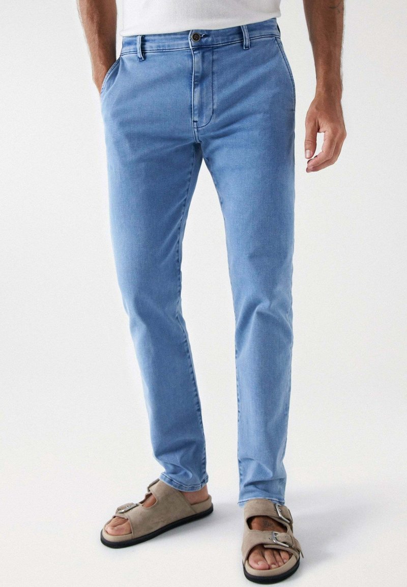 Homme portant un jean slim bleu clair et des sandales beiges à orteils ouverts avec des brides à boucle, debout devant un fond uni.