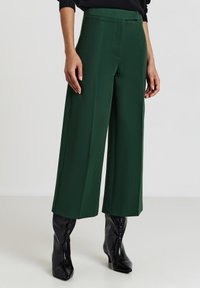 Pantaloni larghi verdi realizzati in tessuto testurizzato, con vita alta e dettagli delle pieghe, abbinati a stivali neri con punta affusolata.
