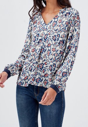 Vrouw draagt een bloes met bloemenprint, lange mouwen, een V-hals en marineblauwe manchetten, gecombineerd met donkerblauwe denim jeans.