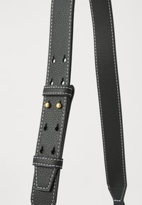 Μαύρος υφασμάτινος δερμάτινος ιμάντας με λευκή ραφή, διαθέτοντας ρυθμιζόμενες τρύπες και αξεσουάρ σε χρυσό τόνο. Κομψός, μινιμαλιστικός σχεδιασμός.