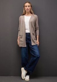 Tansprucket stickad cardigan med fickor, vit skjorta och vida blå jeans. Modellen bär ljusa sneakers. Slät textur, avslappnad passform.